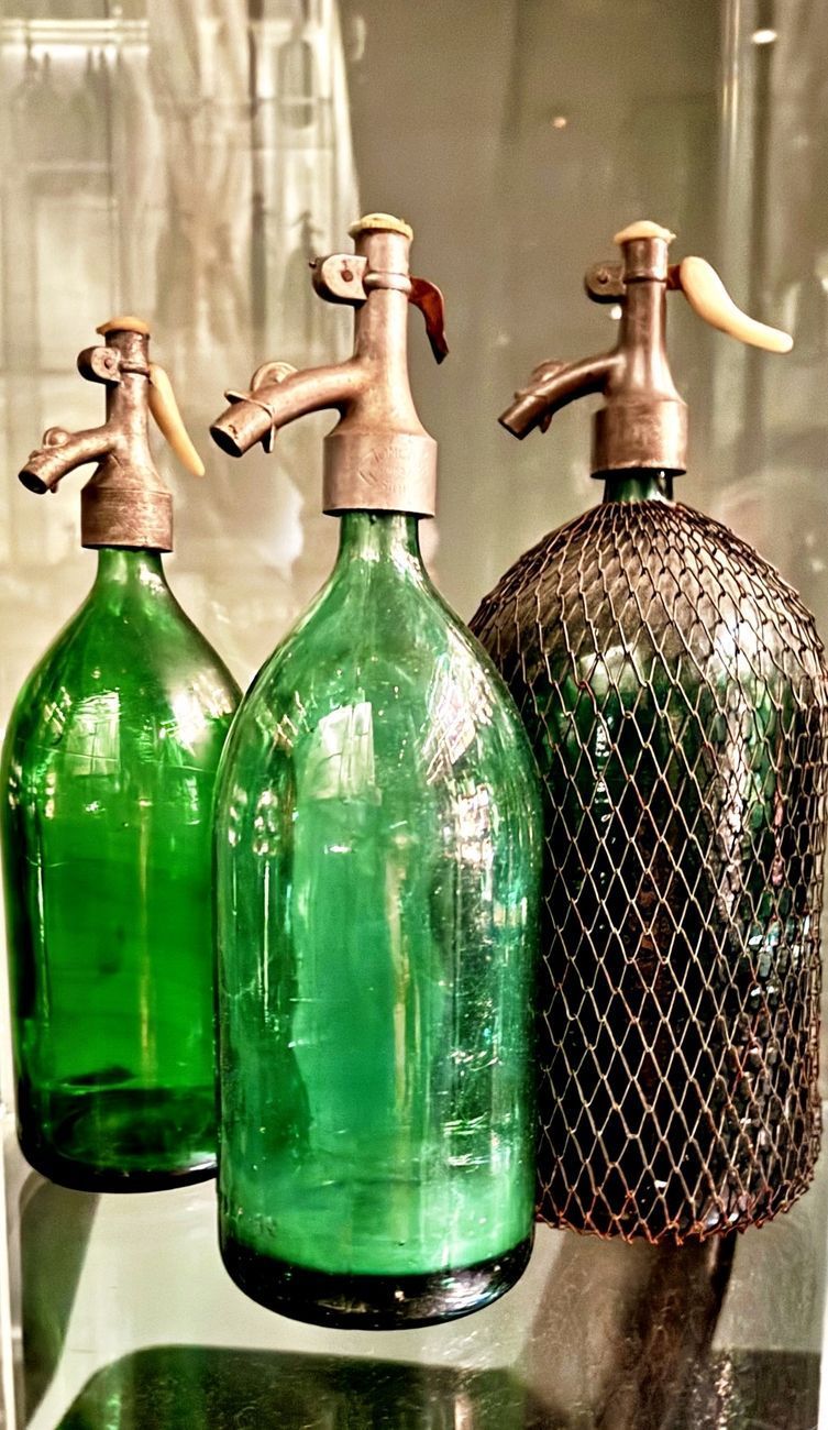 Three vintage green glass soda siphons with brass spouts, one in a wire mesh.