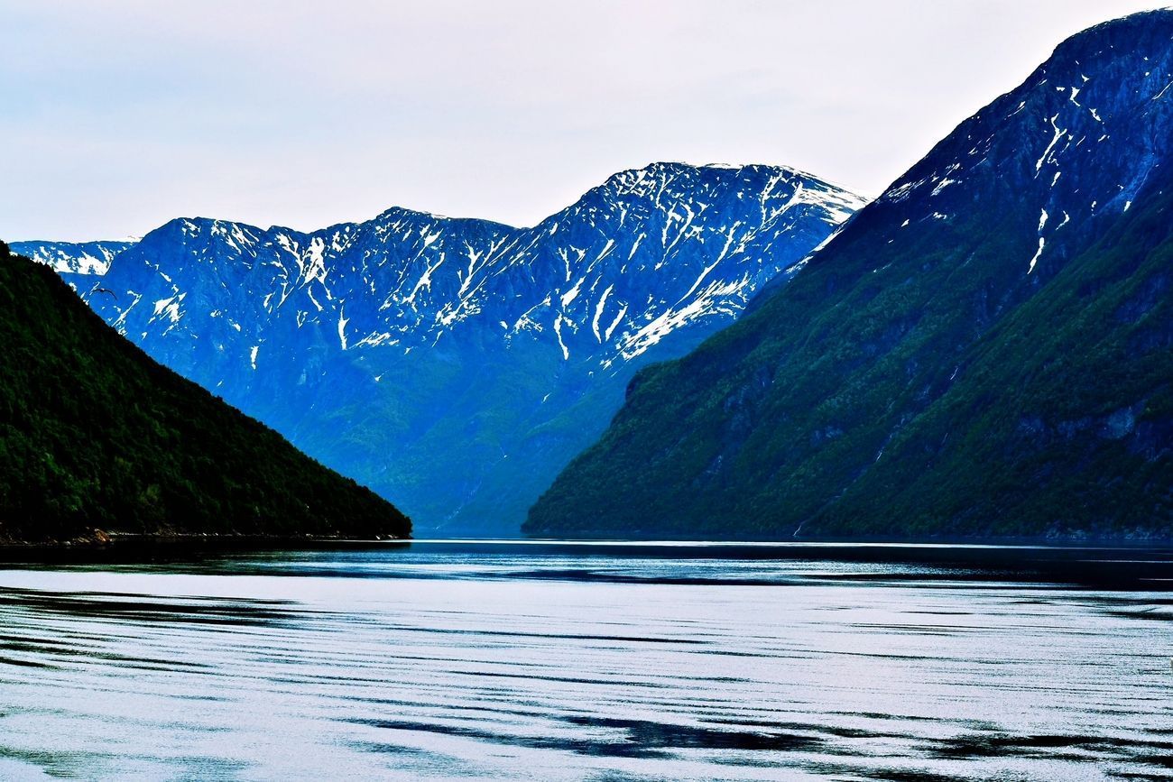 Fjord with dark water, flanked by lush green cliffs and snowy, blue-toned mountains under a cloudy sky.