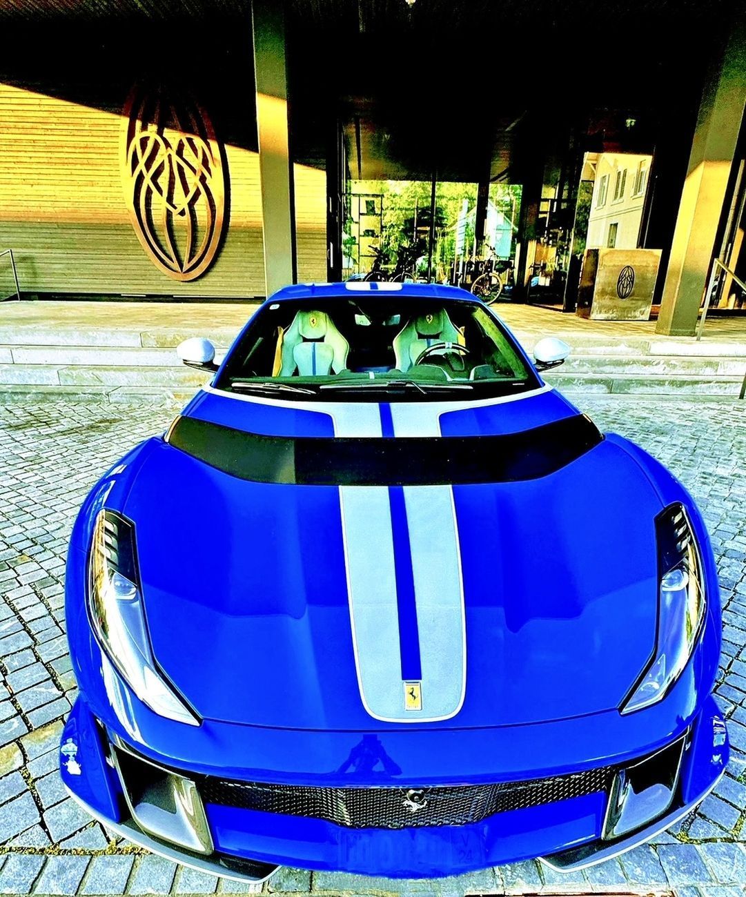 Blue Ferrari sports car with white racing stripes parked in front of a modern building.