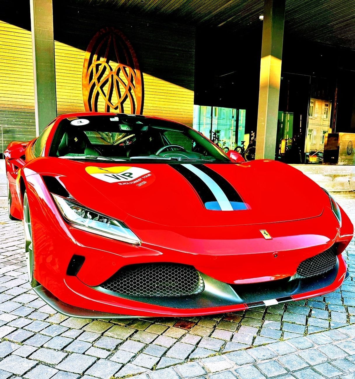 Red Ferrari sports car with black and blue racing stripes parked on cobblestone.