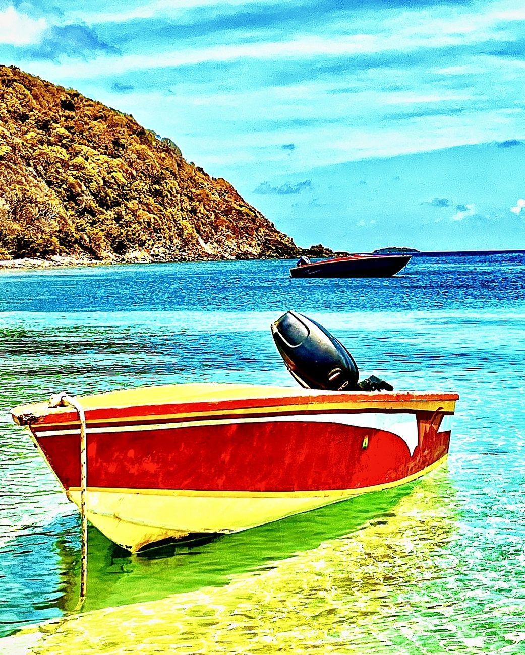 Red and yellow boat floats on turquoise water near a mountain and another boat.
