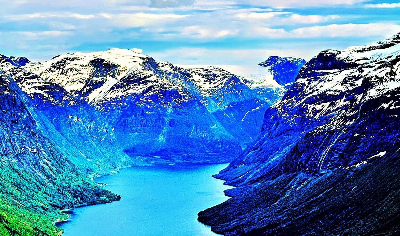 Snow-capped mountains flank a bright blue fjord. Green forest meets the water.