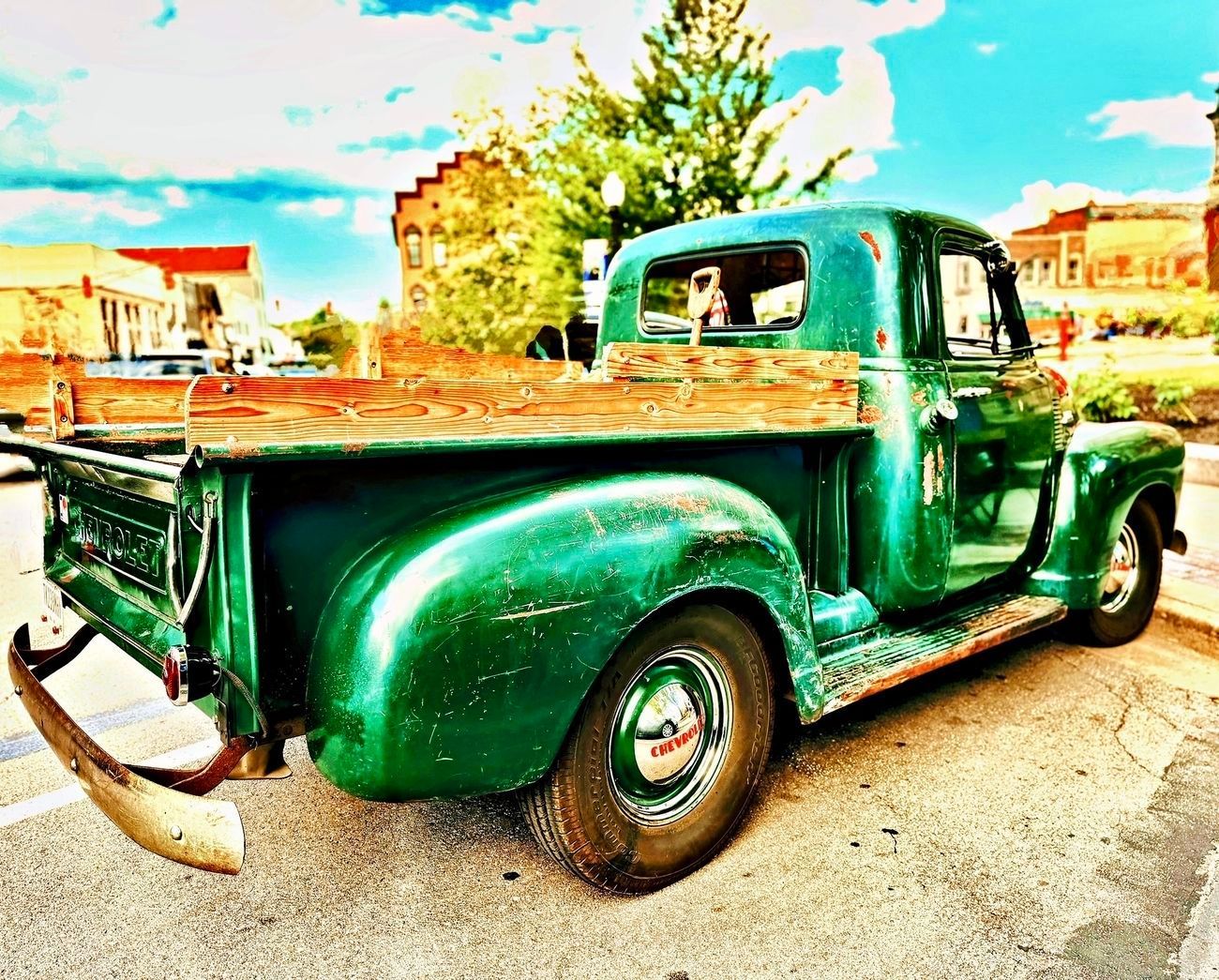 Green vintage pickup truck parked on street, wooden bed, buildings in the background.