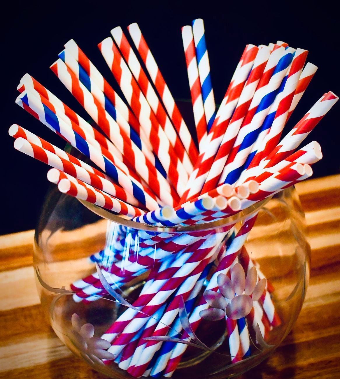 Red, white, and blue striped paper straws in a glass bowl on a wooden surface.