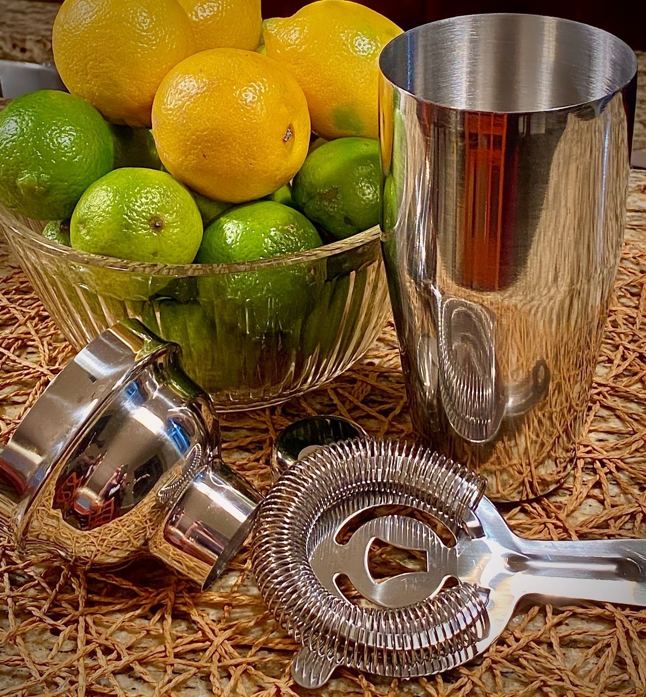 A cocktail shaker, strainer, and bowl of lemons and limes on a woven mat.