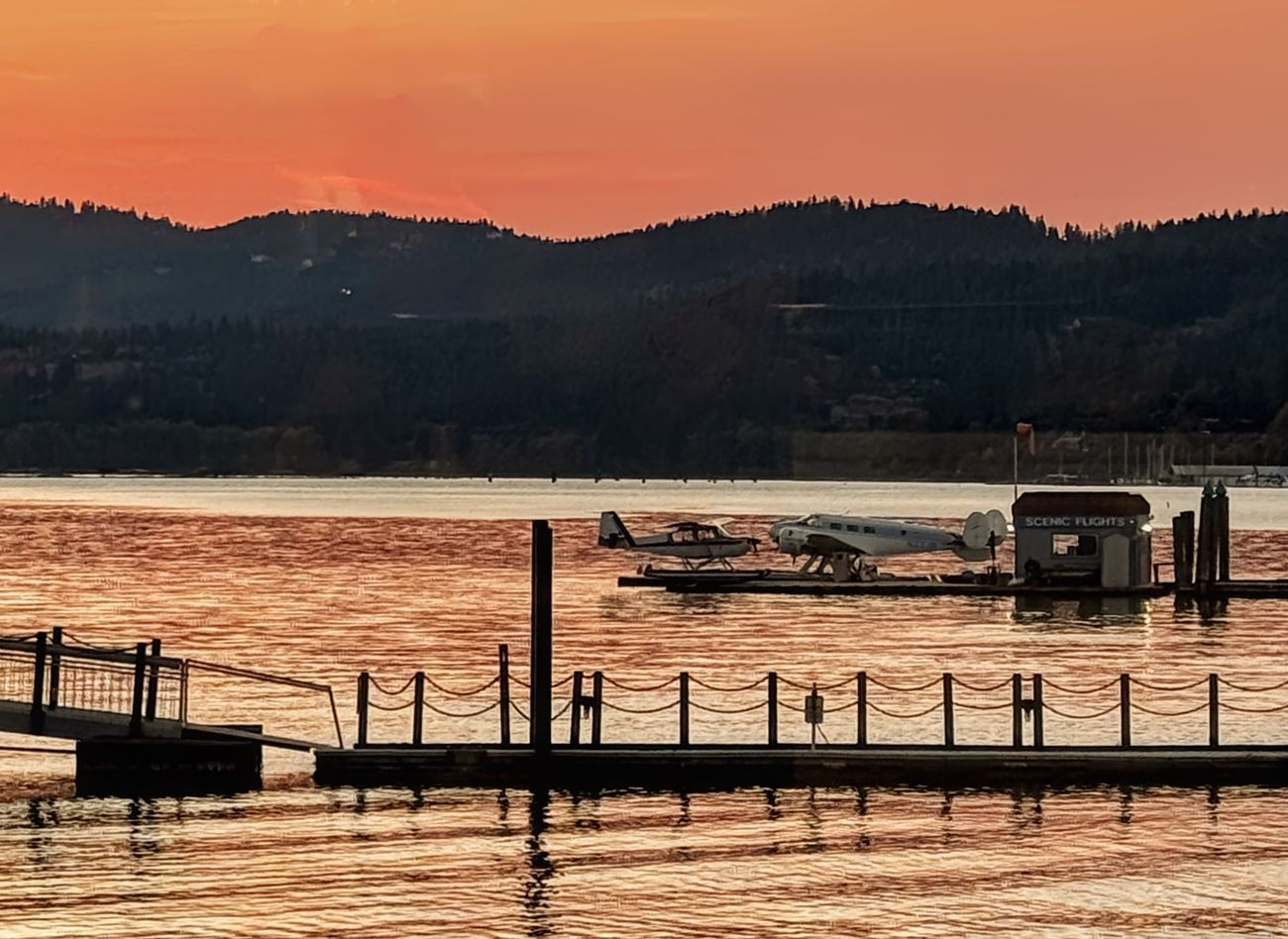 Sunset over lake with seaplanes docked at pier; mountains in the background.
