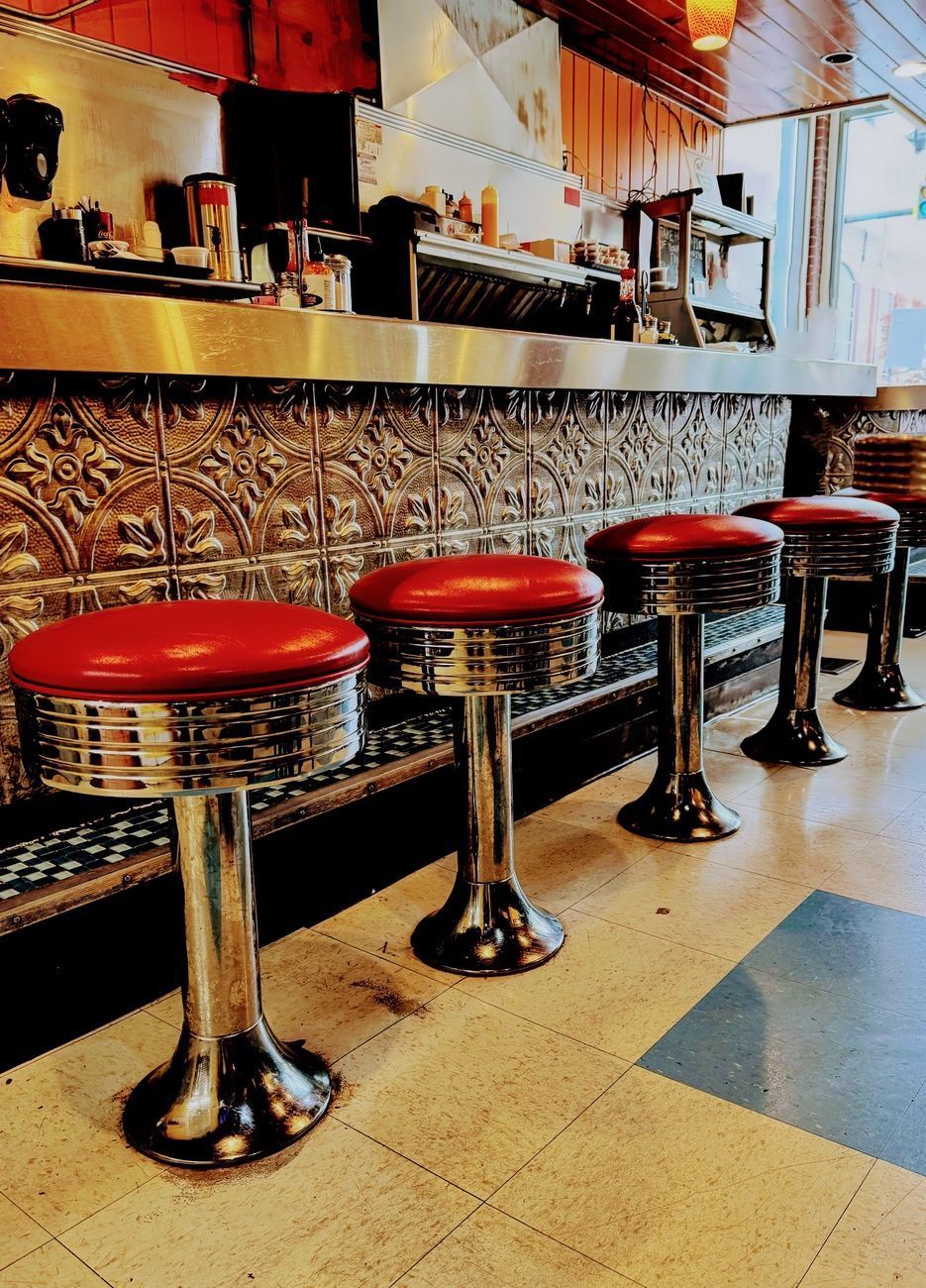 Row of red diner stools at a counter, with metal detailing and patterned wall.