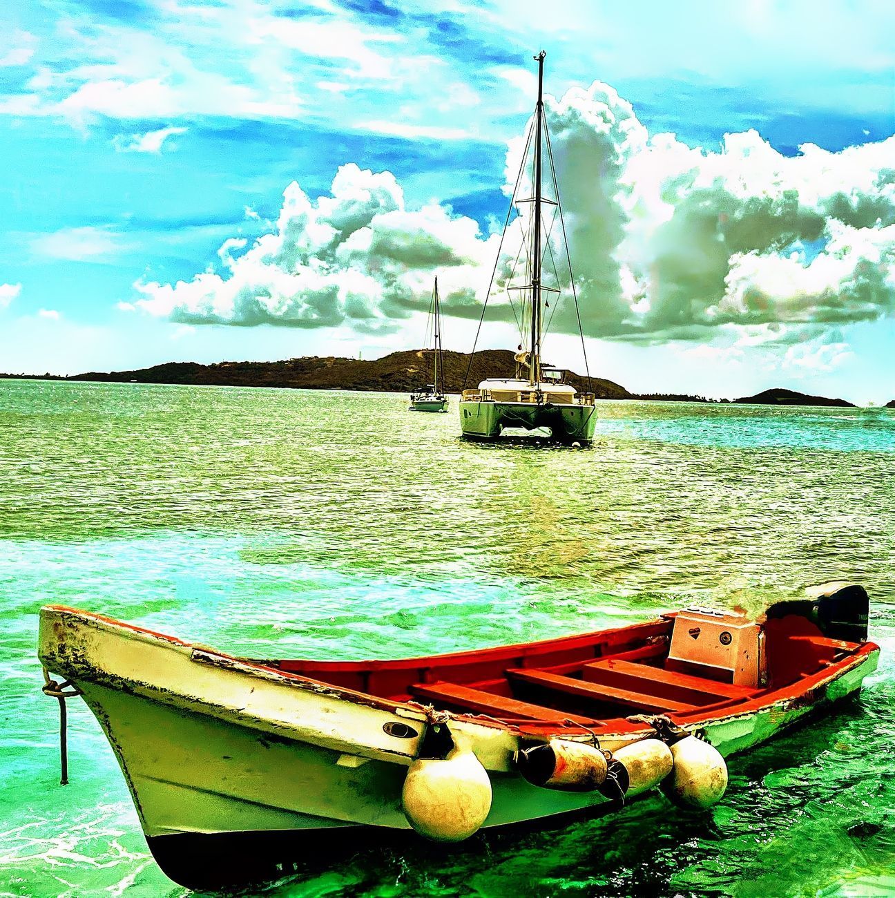 Small, colorful boat floats on turquoise water; sailboat in background under a cloudy blue sky.