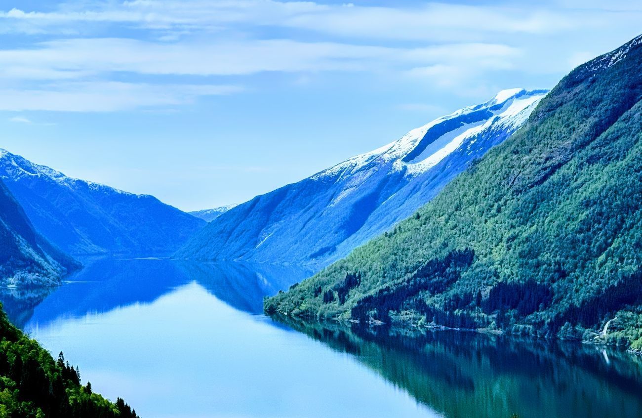 A tranquil fjord surrounded by mountains with patches of snow reflecting in the calm water.