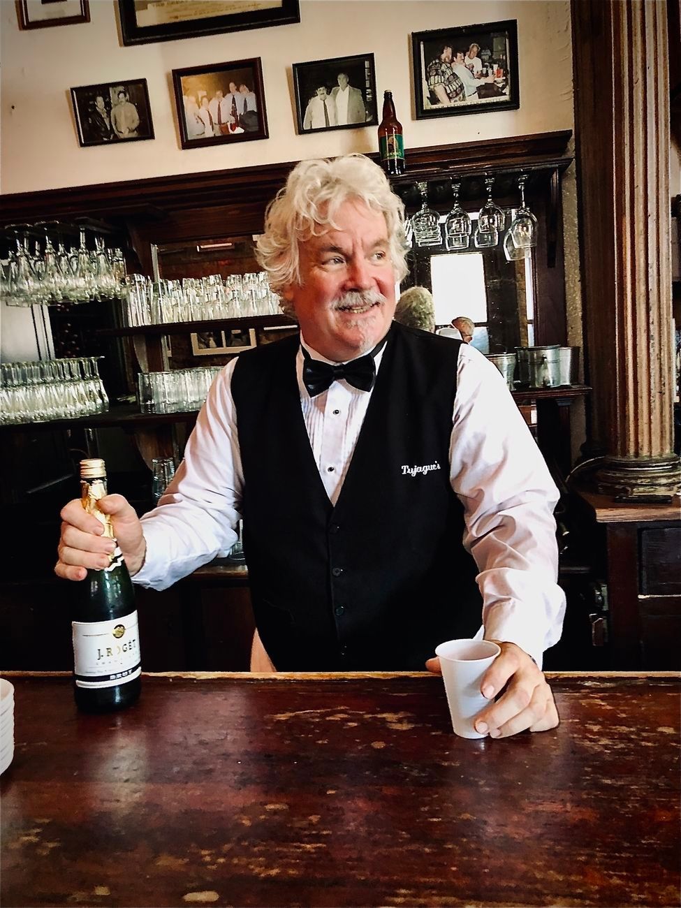 Bartender holding champagne bottle and cup behind a wooden bar.