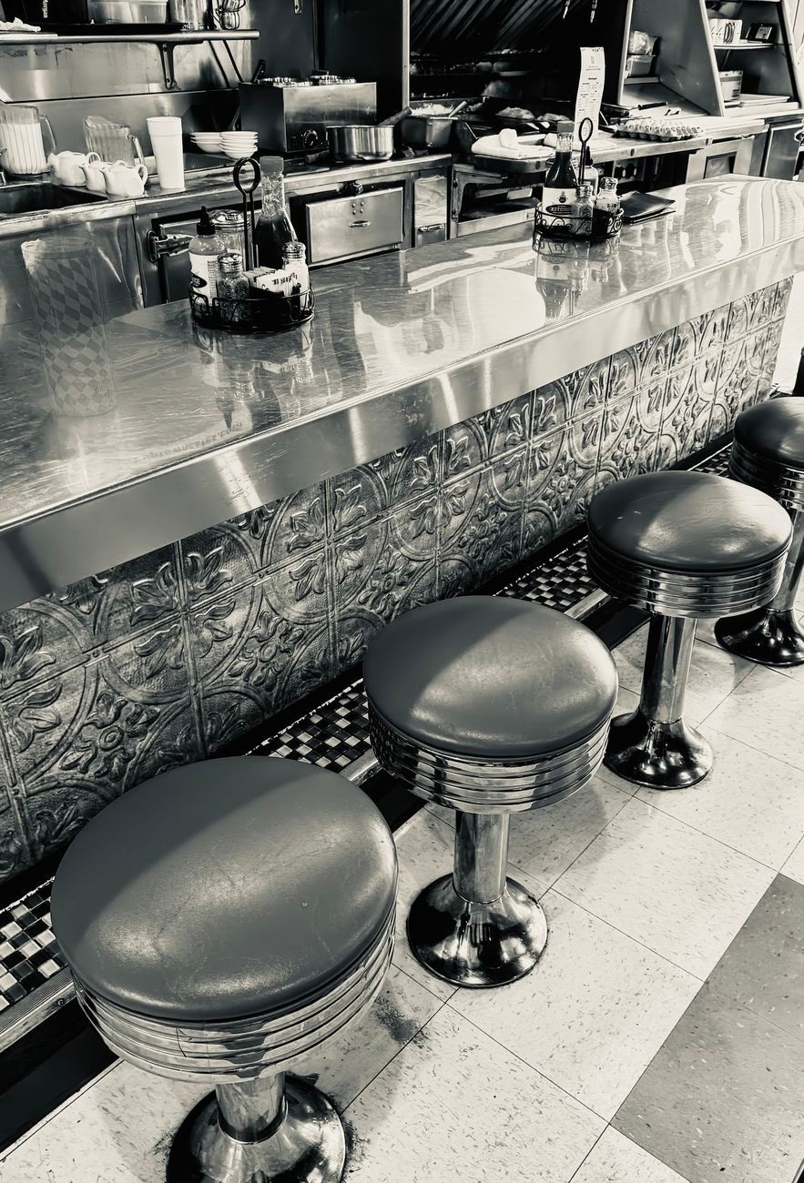 Black and white photo of an empty diner counter with chrome stools.