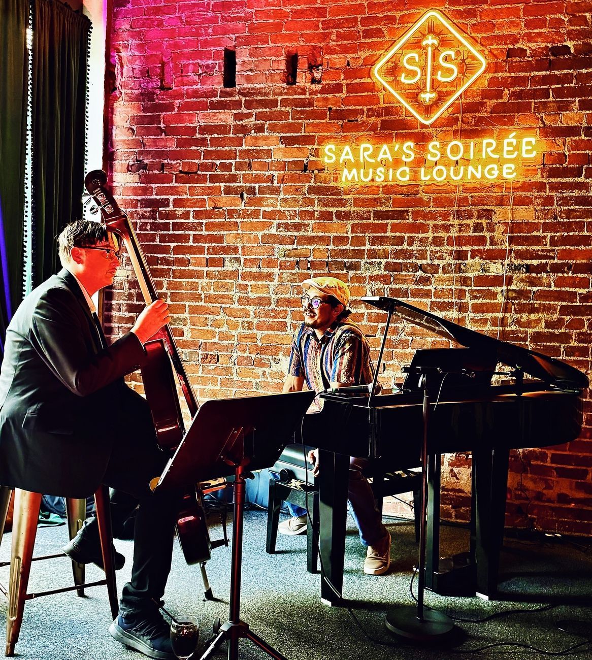 Jazz musicians perform at Sara's Soirée Music Lounge. A bassist and pianist play in front of a brick wall and neon sign.