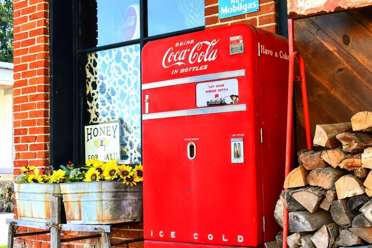 Red vintage Coca-Cola vending machine outside a brick building, sunflowers in metal tubs.