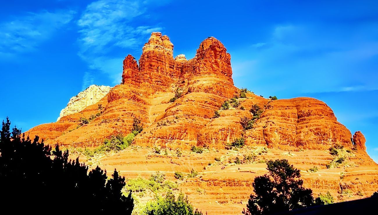 Red rock butte against a bright blue sky with sparse greenery at the base.
