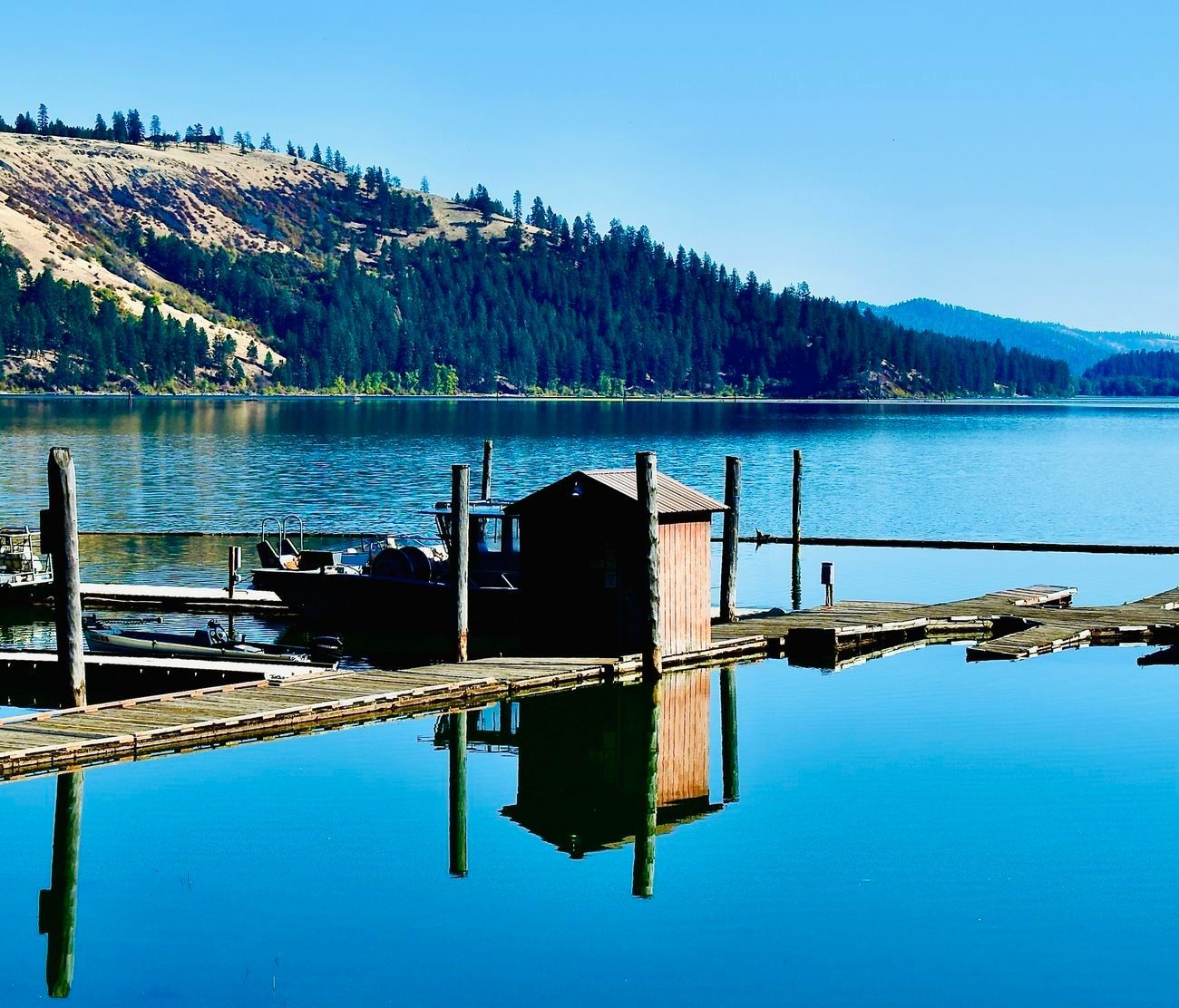 Wooden dock and shed on calm, blue lake with forested hills in the background under a clear sky.