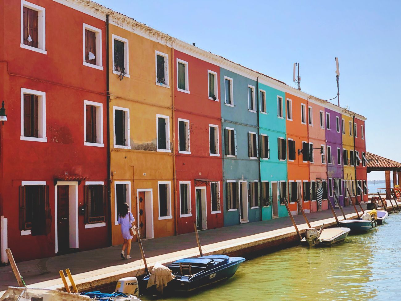 Colorful row of houses along a canal, Burano, Italy. A person walks by; boats are moored.