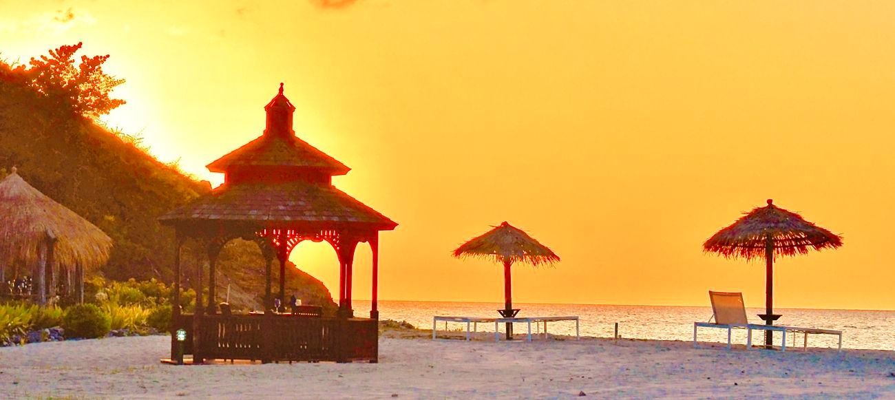 Beach scene at sunset with gazebo, umbrellas, and ocean. Orange sky, golden light.