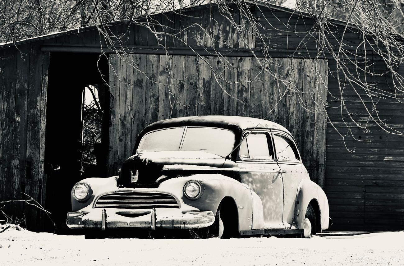 Vintage car covered in snow in front of a wooden shed in black and white.