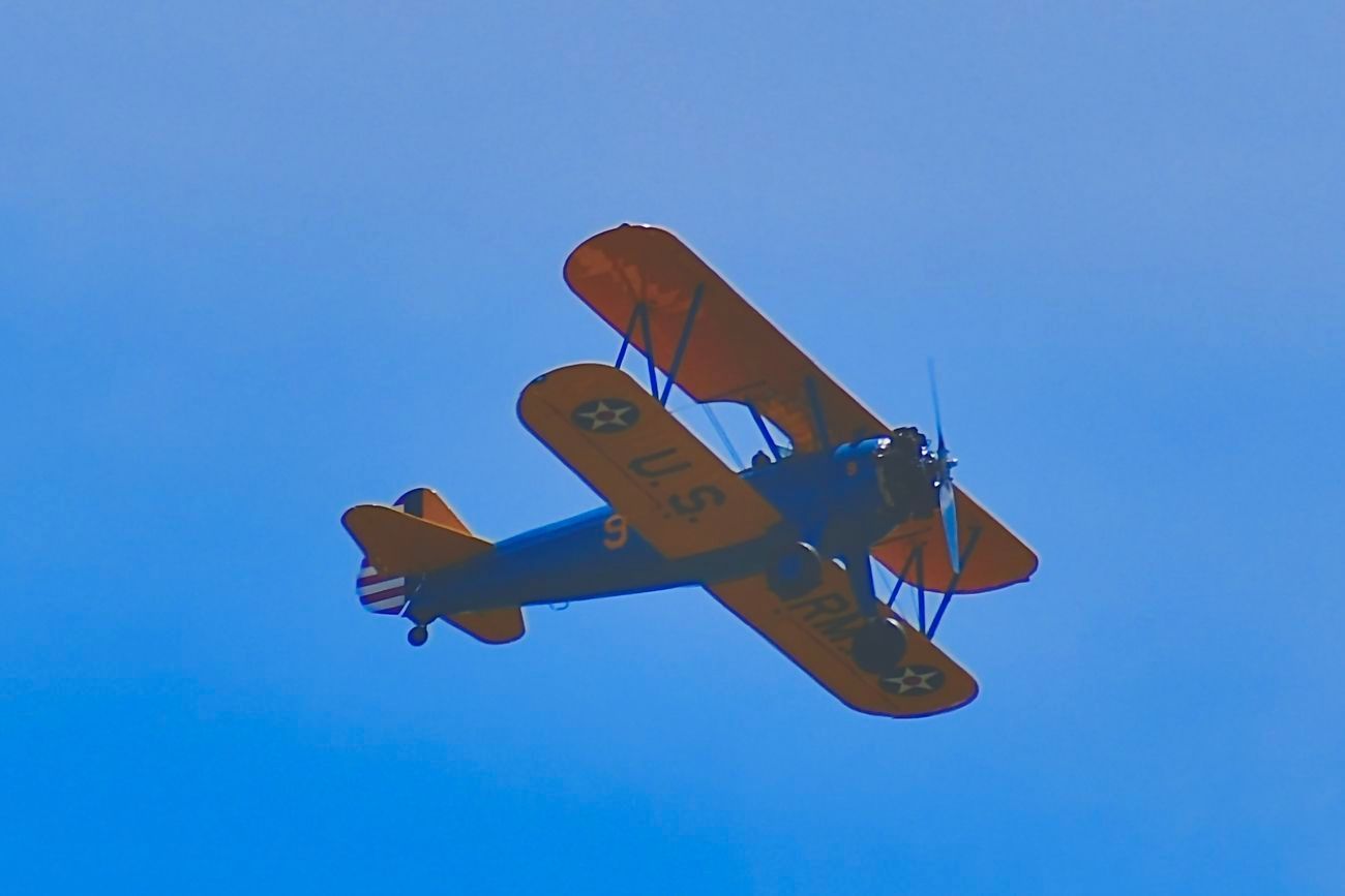 Biplane with yellow wings and blue fuselage flying in a clear blue sky.