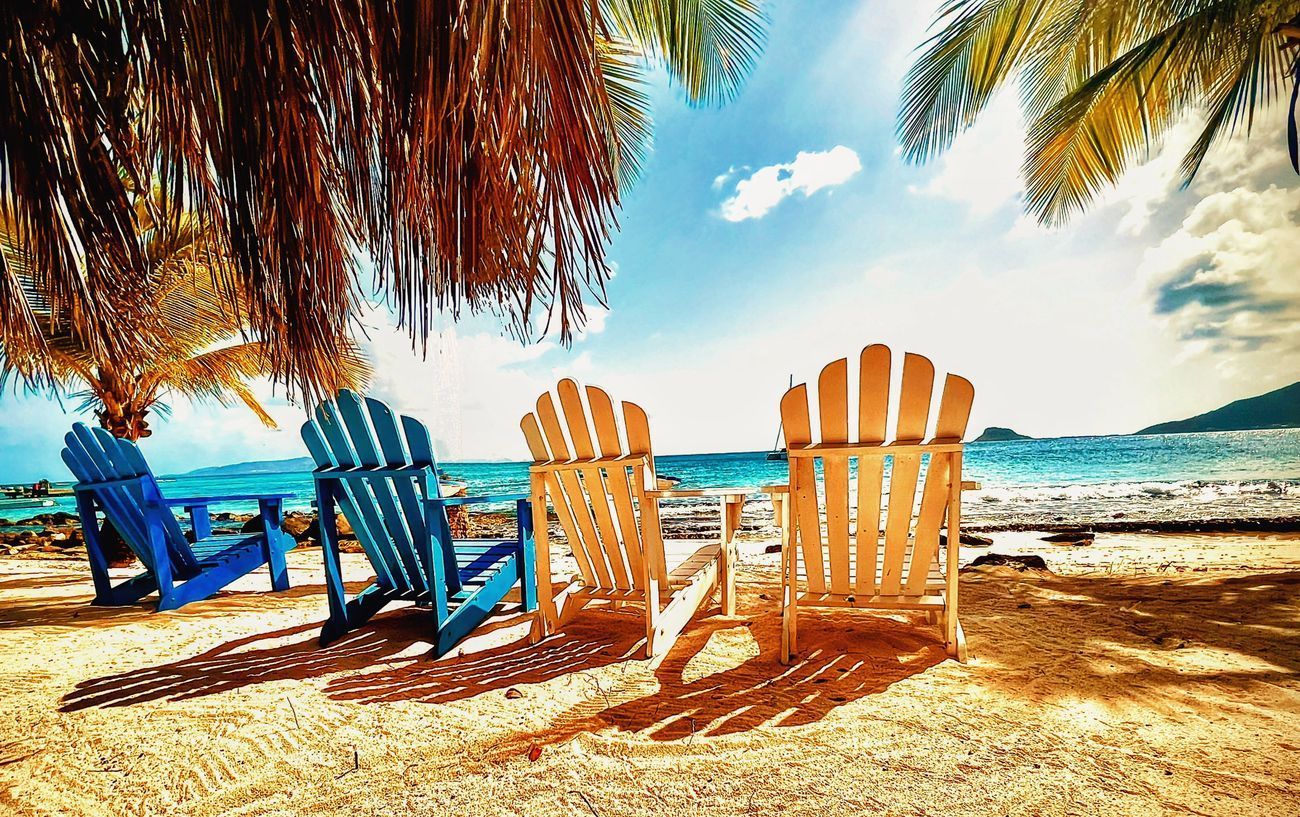 Three Adirondack chairs on a sandy beach under palm trees, overlooking a blue ocean.