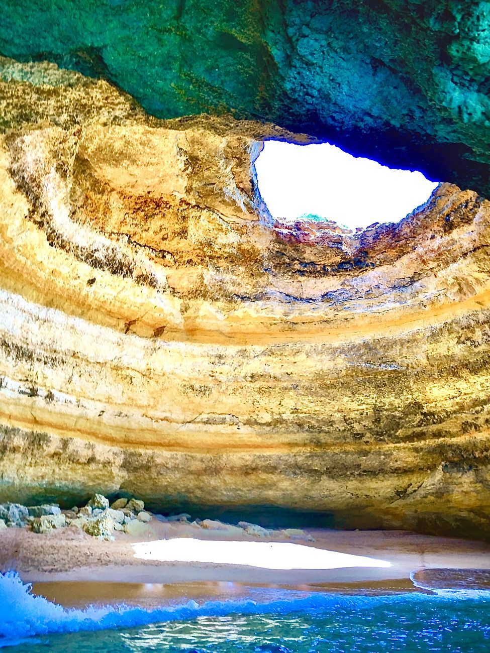 Cave interior with a circular opening, sandy beach, and turquoise water.