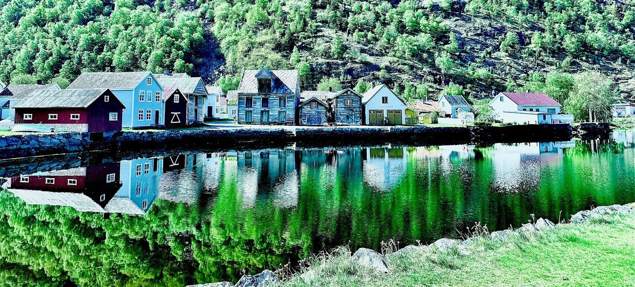 Colorful buildings along a calm body of water with reflections of the buildings and trees. Lush green hillside in the background.