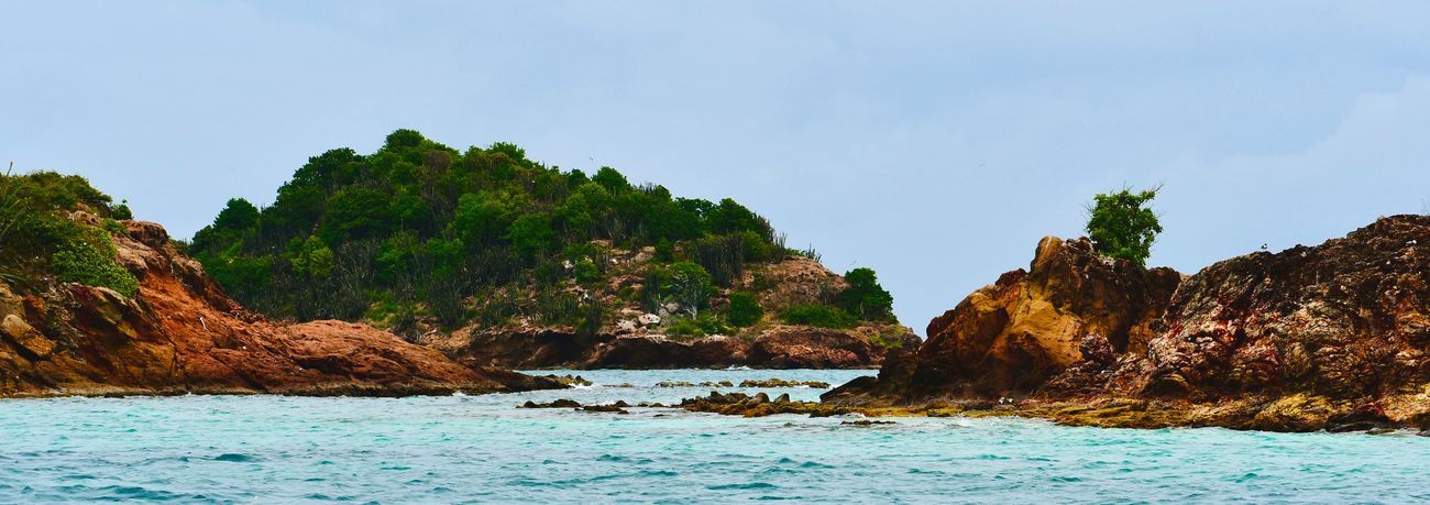 Island with lush greenery, rocky shores, and blue water under a cloudy sky.