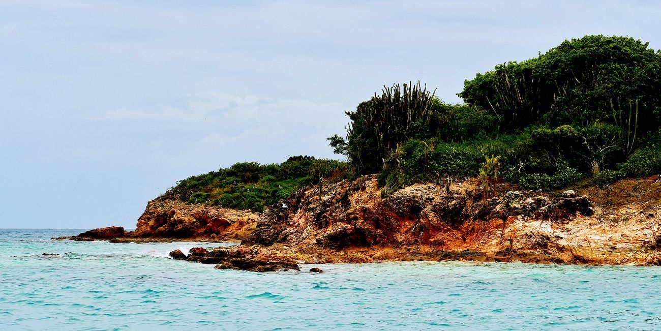 Coastal view of an island with lush green vegetation, brown rocky cliffs, and turquoise water under a cloudy sky.