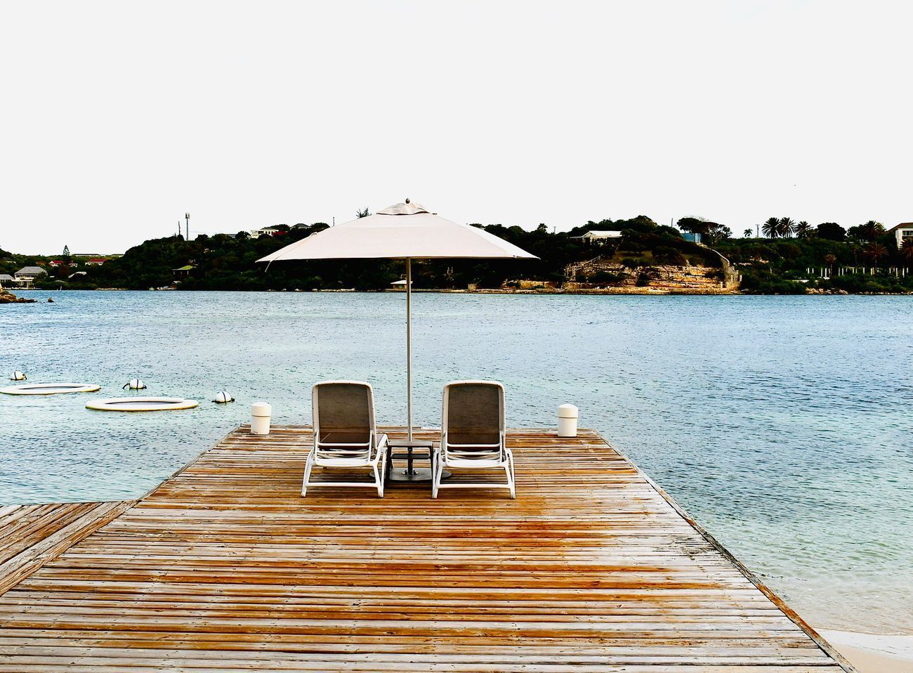 Two lounge chairs under a white umbrella on a wooden dock overlooking calm water.