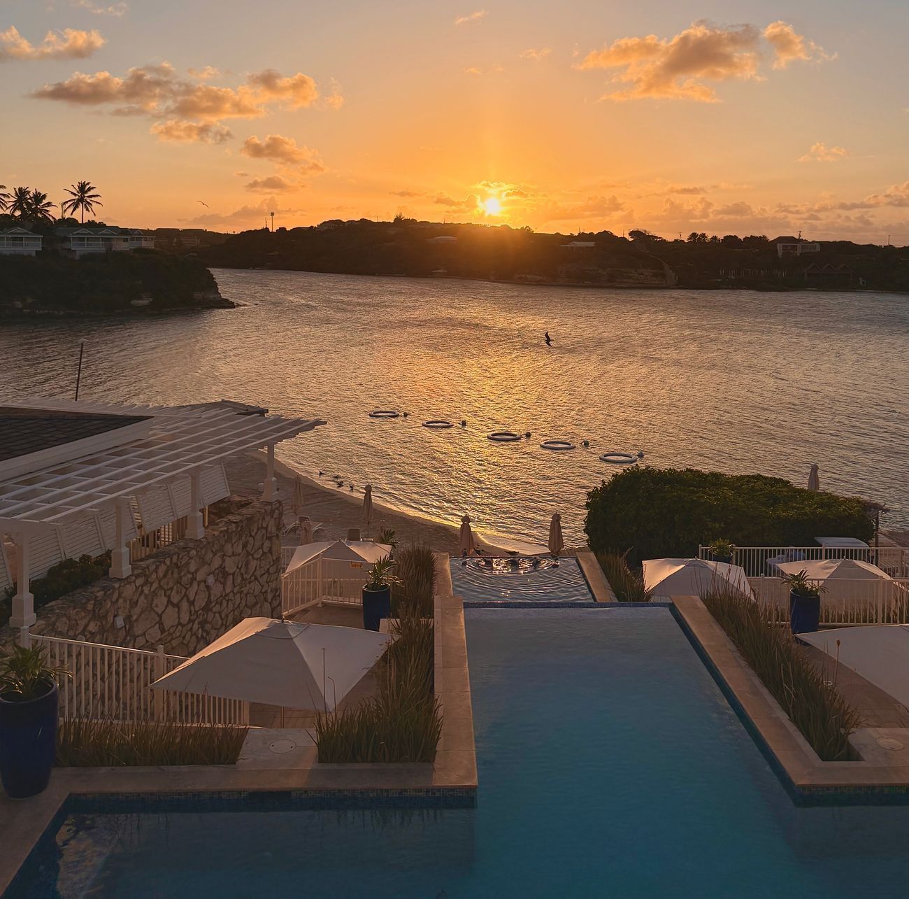 Sunset over a body of water, with a pool and white cabanas in the foreground.