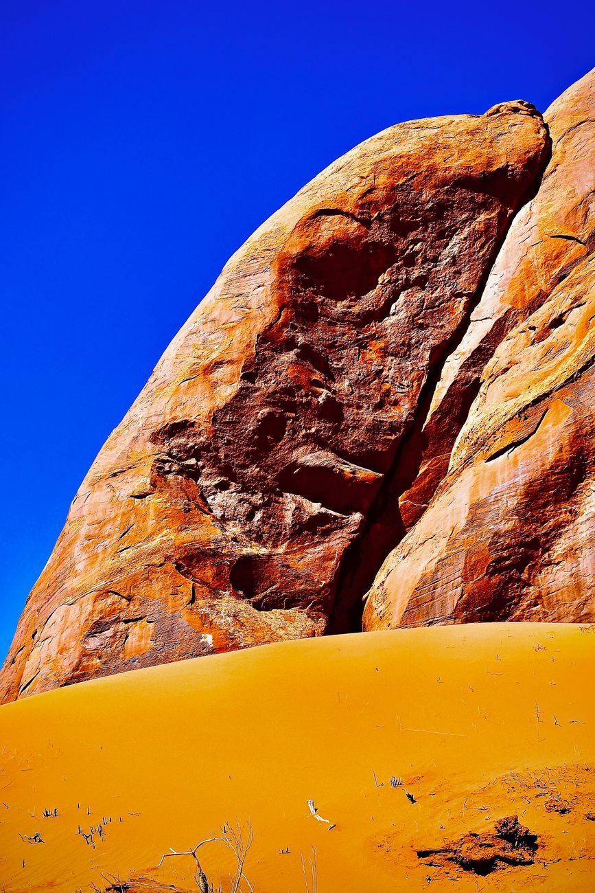Orange rock formation against a blue sky, with a sand dune in the foreground.