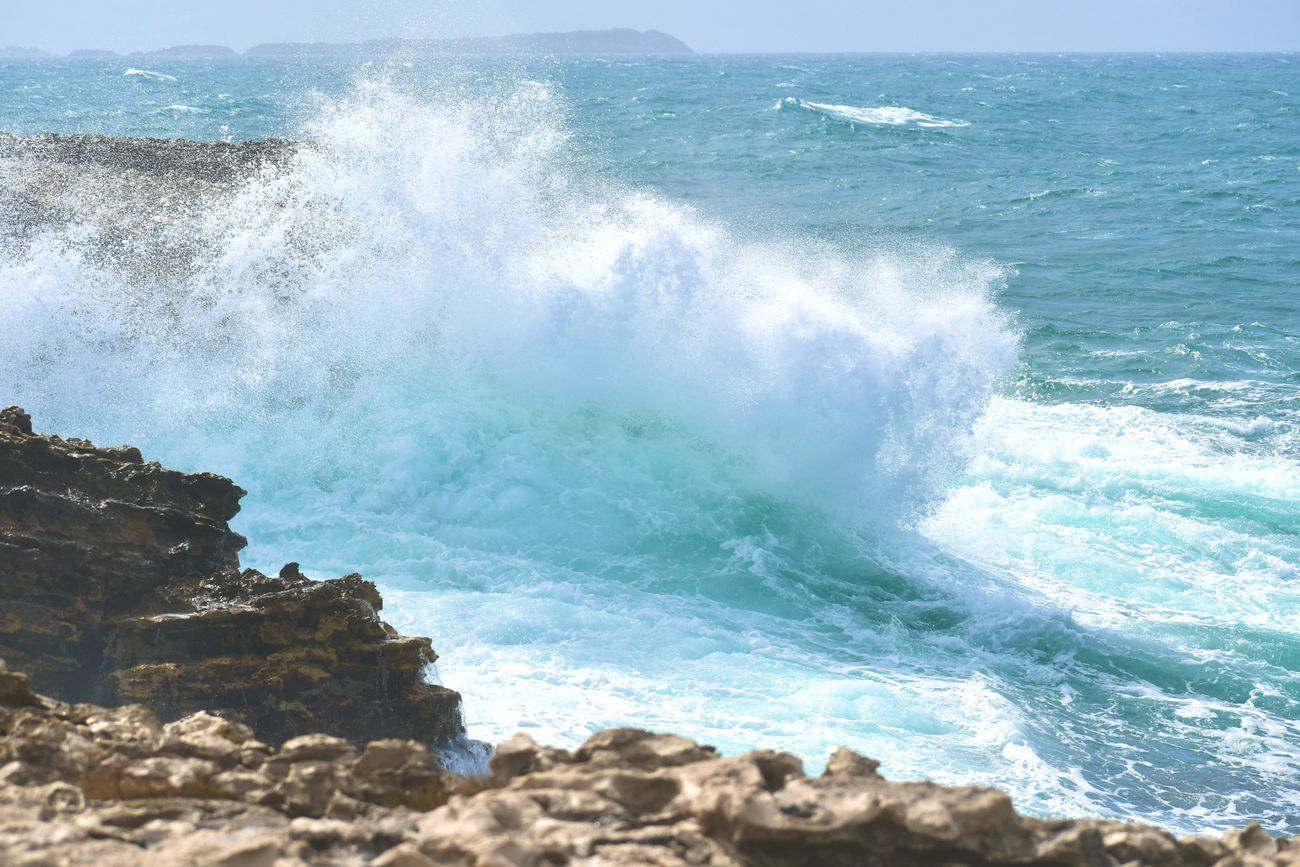 Ocean wave crashing against rocky shore, spraying white foam and turquoise water.