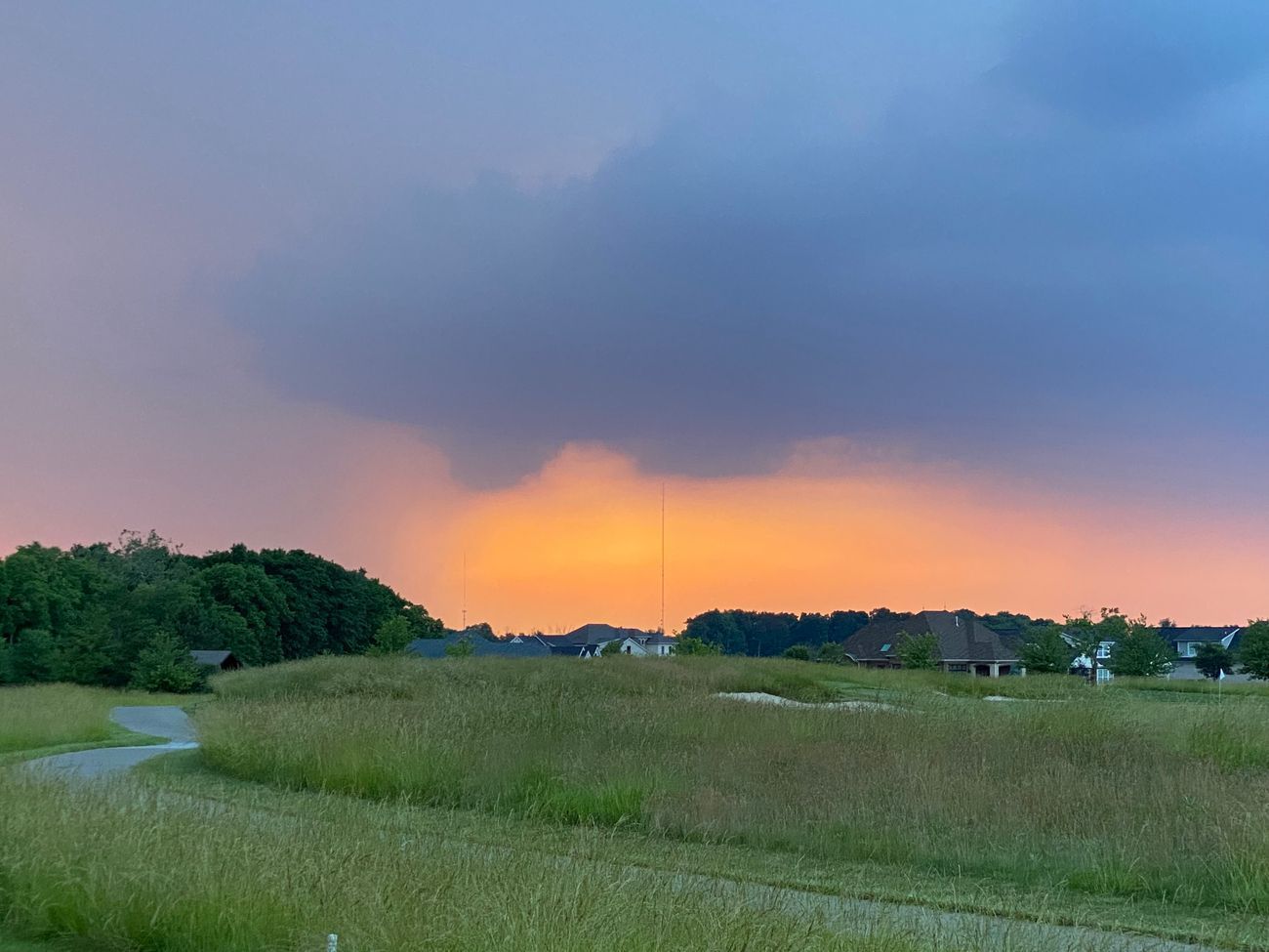 Grassy field with a walking path, trees, and houses under a colorful sunset sky.