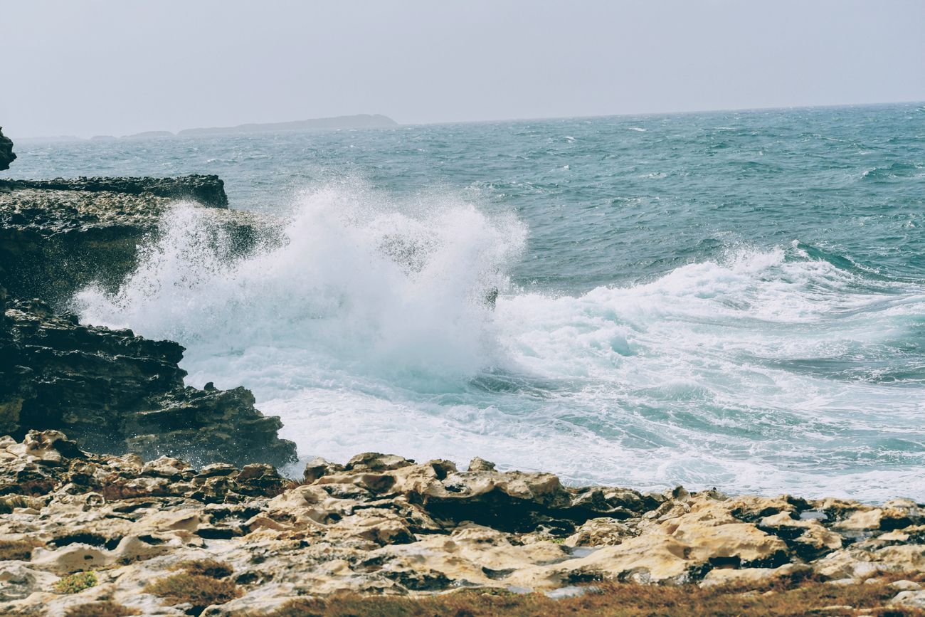 Ocean waves crashing against rocky shoreline. White foam, blue water, and sunlit rocks.