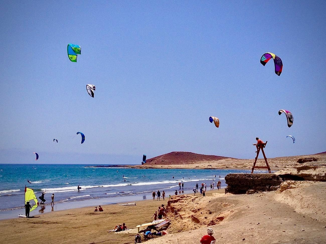 Kitesurfers on a sunny beach with blue water and sky; several colorful kites in the air.