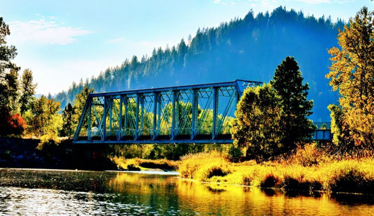 Bridge over a river, surrounded by trees and a mountain, under a blue sky.