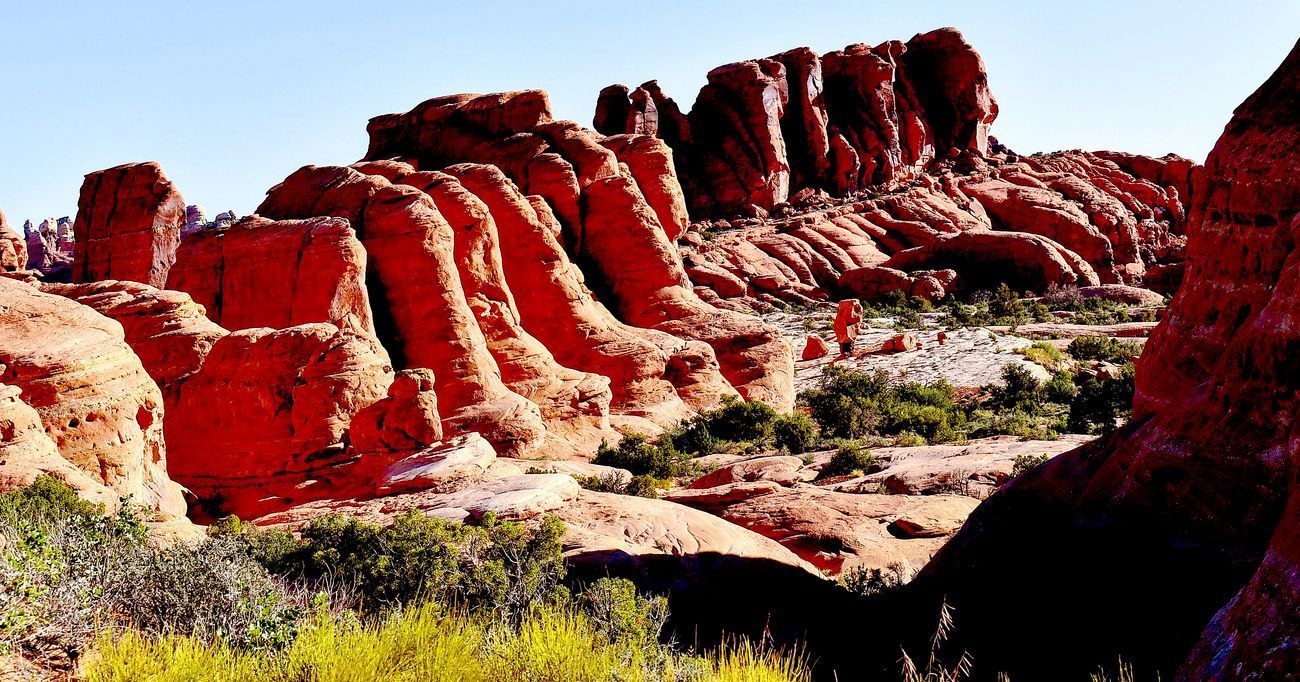 Red rock formations under a clear blue sky, with sparse green vegetation in a desert landscape.