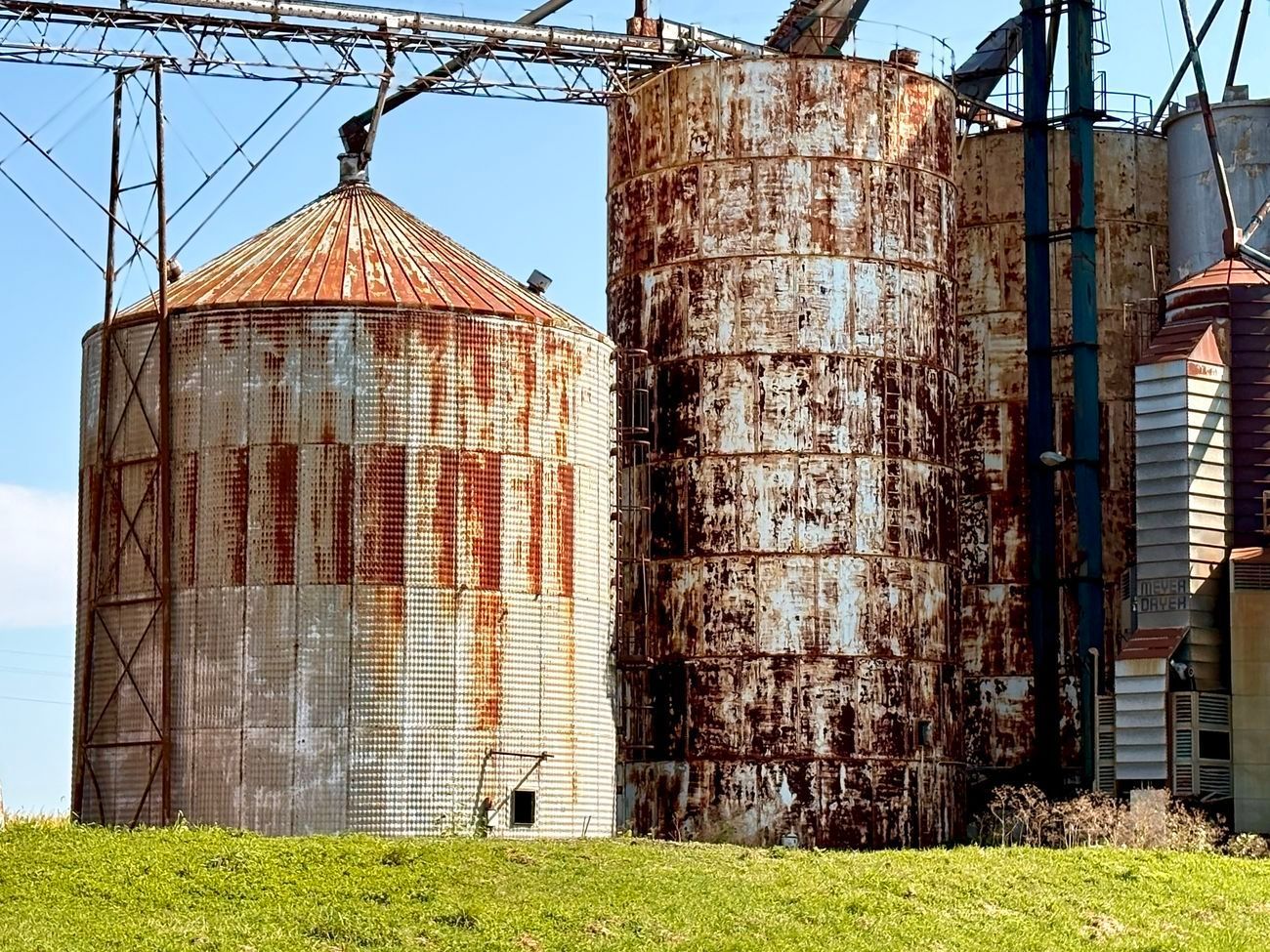 Rusty grain silos against a blue sky, standing on a grassy hill.