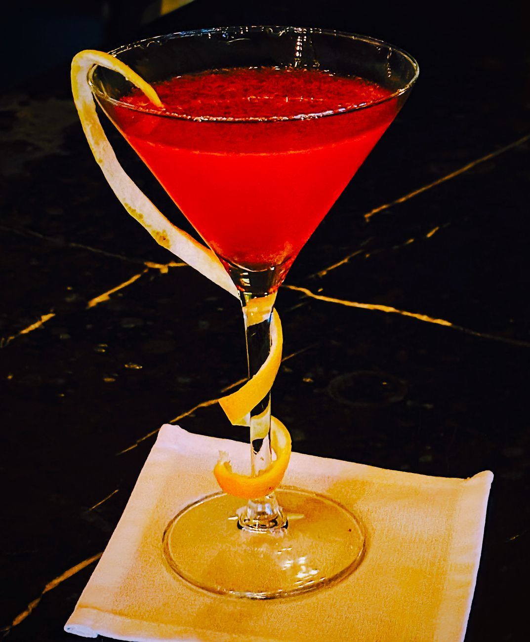 Red cocktail in a stemmed glass with a lemon twist on a white napkin.
