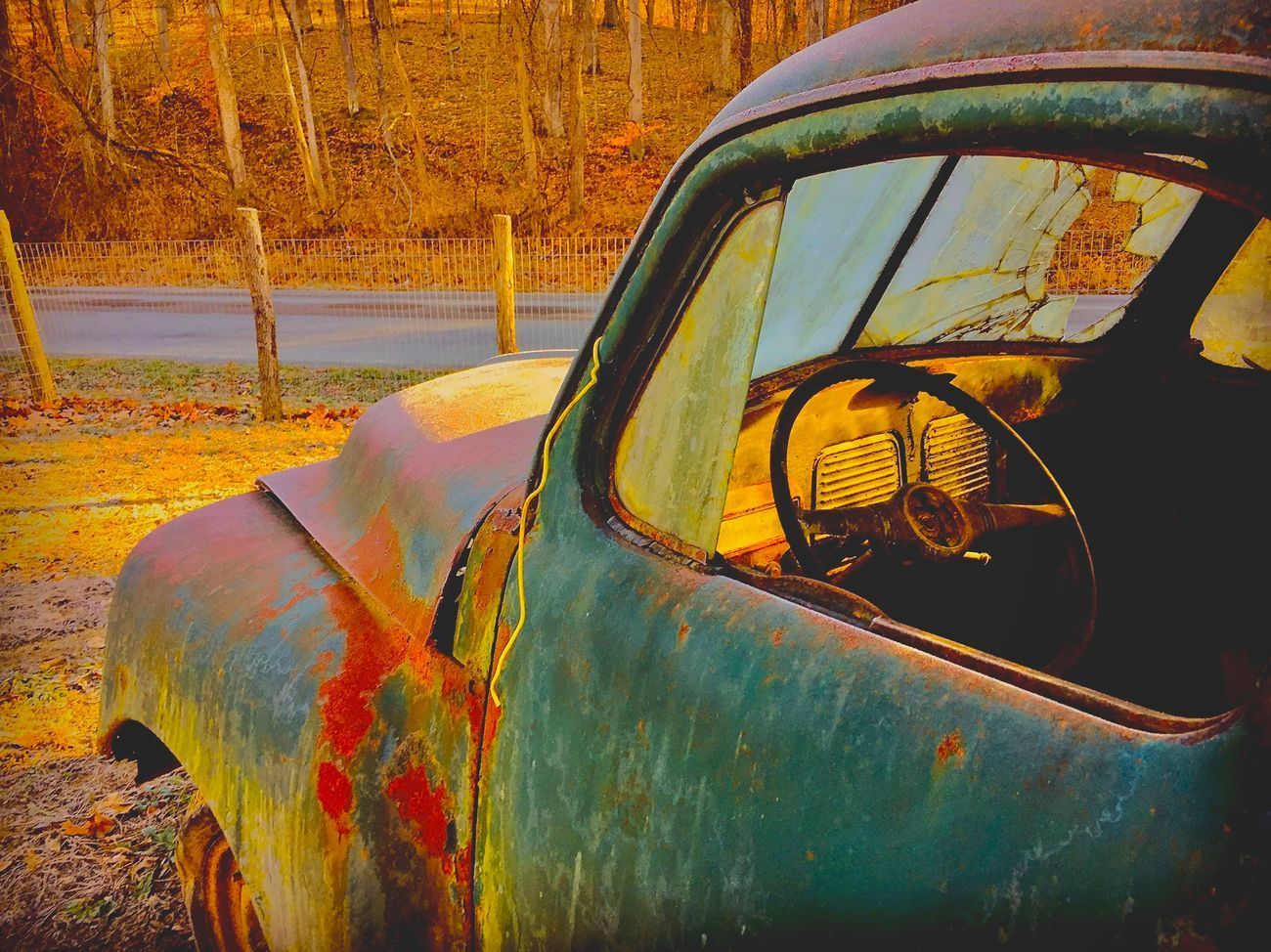 Rusty blue and red vintage pickup truck, outdoors. Wooden fence and trees in background.