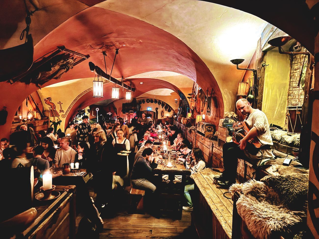 Dimly lit, crowded restaurant interior; people dining at tables, man playing guitar on the right.