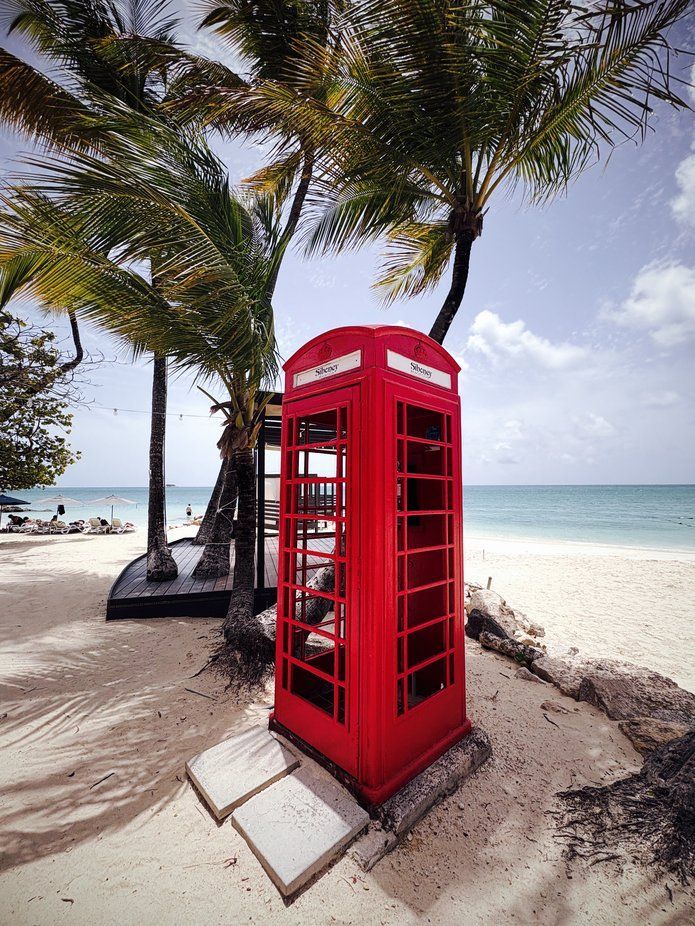 Red British phone booth on a sandy beach under palm trees, with the ocean in the background.
