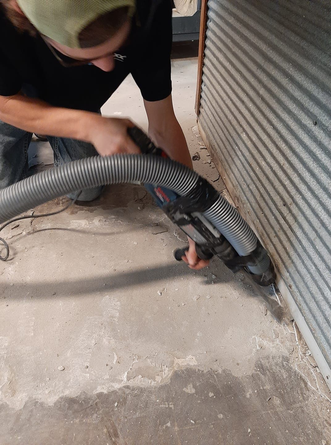 A person uses a power tool with a vacuum attachment to cut along the edge of a concrete floor against a metal wall.