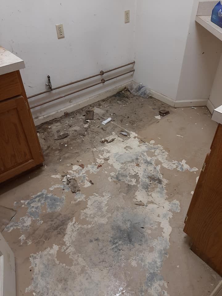 An unfinished, cluttered room with bare concrete floors showing adhesive remnants and exposed wall pipes.