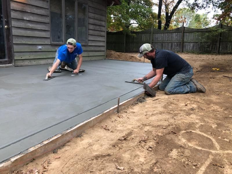 Two workers kneel on a freshly poured concrete patio, smoothing the surface with hand trowels alongside a wooden home.