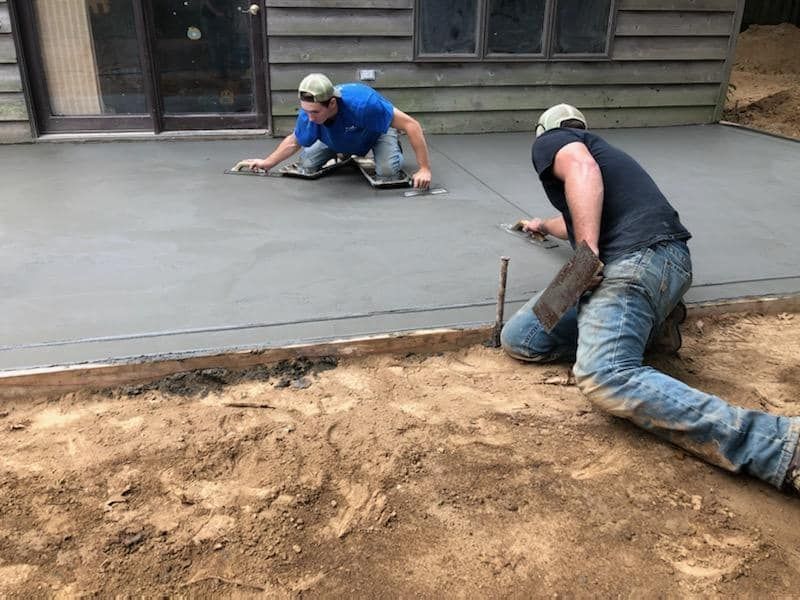 Two workers kneel on a freshly poured concrete patio, smoothing the surface with hand tools next to a house.