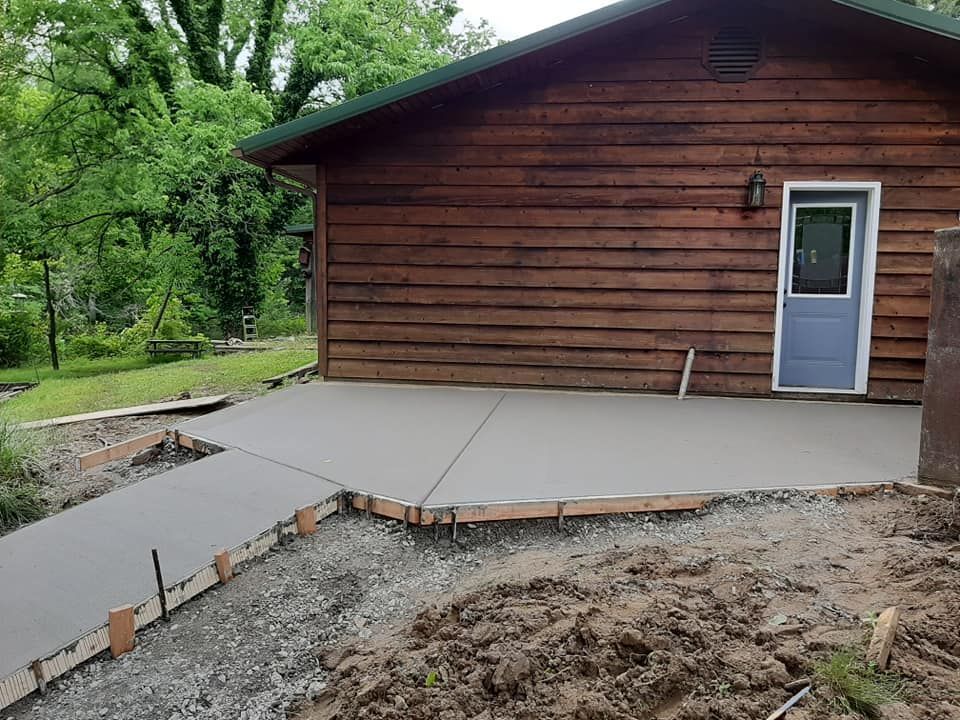 A new, gray concrete patio and walkway extend from a brown wooden cabin with a light blue door set in a grassy yard.