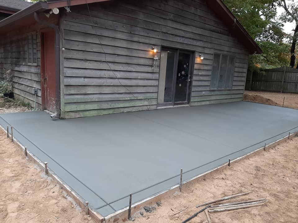 A newly poured gray concrete patio slab next to a rustic, wood-sided cabin.