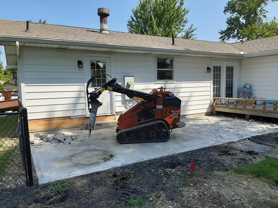 An orange skid-steer loader with a hydraulic breaker attachment sits on a concrete patio beside a white house.