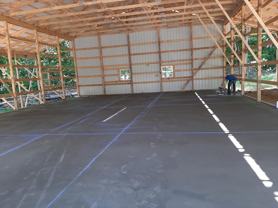 A person stands inside a large, unfinished wooden frame building with a newly poured gray concrete floor and chalk lines.